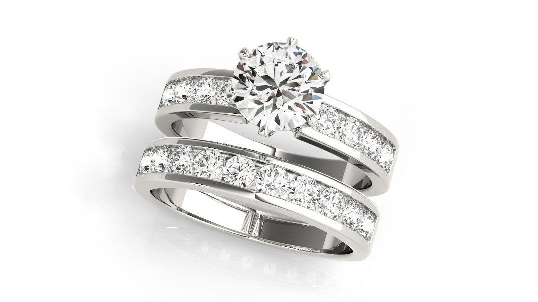 The image shows a luxurious bridal ring set featuring a prominent engagement ring with a large round cut diamond in a prong setting, accompanied by two channel set wedding bands. The bands are crafted in polished white gold, with the wedding bands showcasing a series of smaller, square-cut diamonds set in a channel that spans the top half of each band. The entire set is displayed against a clean, white background, emphasizing the brilliance and clarity of the diamonds.