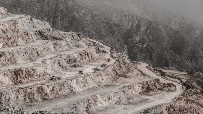 An expansive view of a diamond mine with terraced open-pit excavation. The mine's winding roads are visible, with large mining trucks navigating the rugged terrain. The background shows rocky, barren mountains under a hazy sky.