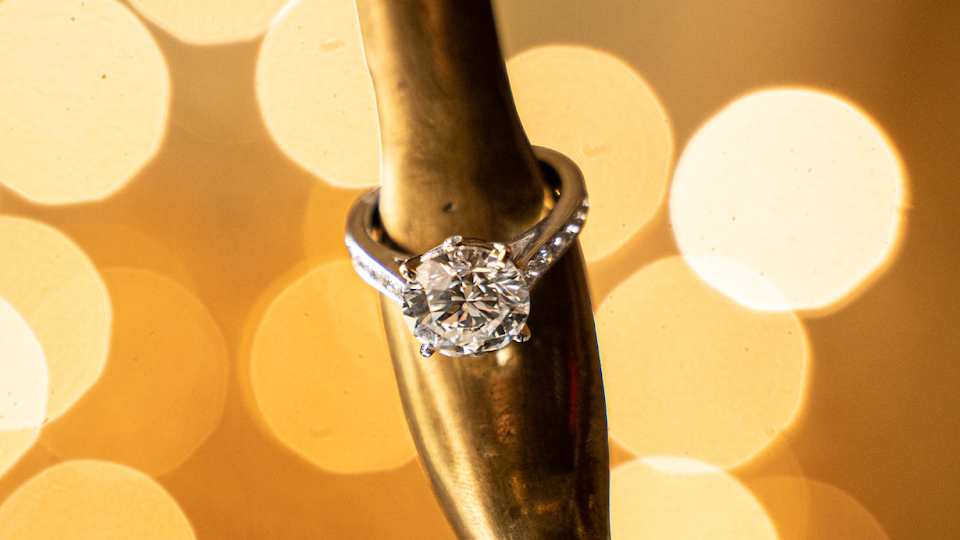 A close-up image of a diamond engagement ring resting on a brass instrument, with a warm bokeh effect of golden lights in the background, creating a romantic and elegant ambiance. The diamond ring is prominently displayed, showcasing its brilliance and clarity