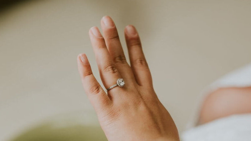 A close-up of a hand with neatly manicured, average-sized fingers wearing a snug-fitting engagement ring. The ring features a prominent oval-shaped diamond center, flanked by smaller diamonds on the band, all sparkling elegantly. The background is blurred, highlighting the ring's beauty